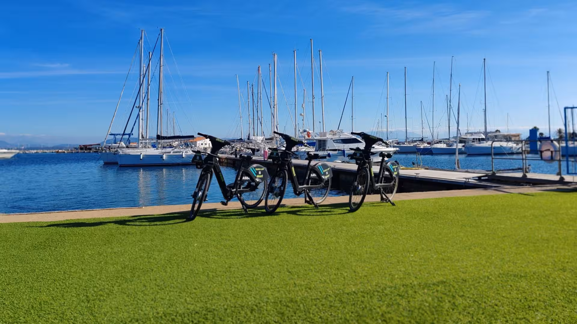Three e-bikes parked on green lawn with sailboats in Calasetta marina under a clear blue sky.