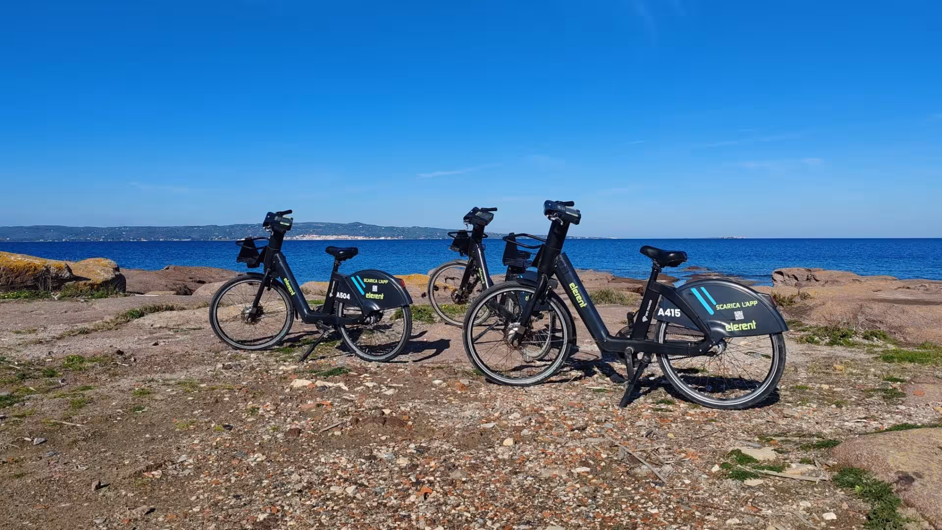 Four e-bikes on rocky shoreline with panoramic sea views in Calasetta, ideal for scenic cycling.