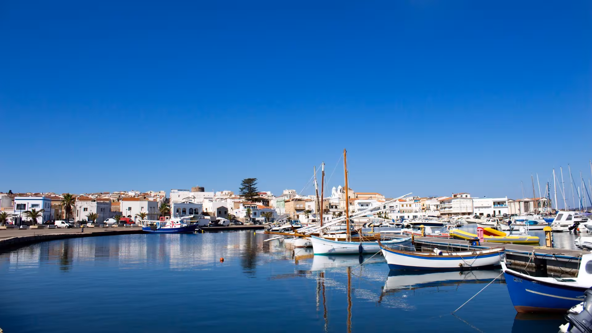 Scenic view of Calasetta harbor with boats docked and whitewashed buildings under clear blue skies.