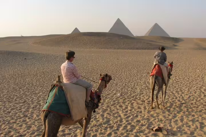 Two riders on camels crossing Giza desert toward pyramids at golden hour, sunrise or sunset tour with transfers
