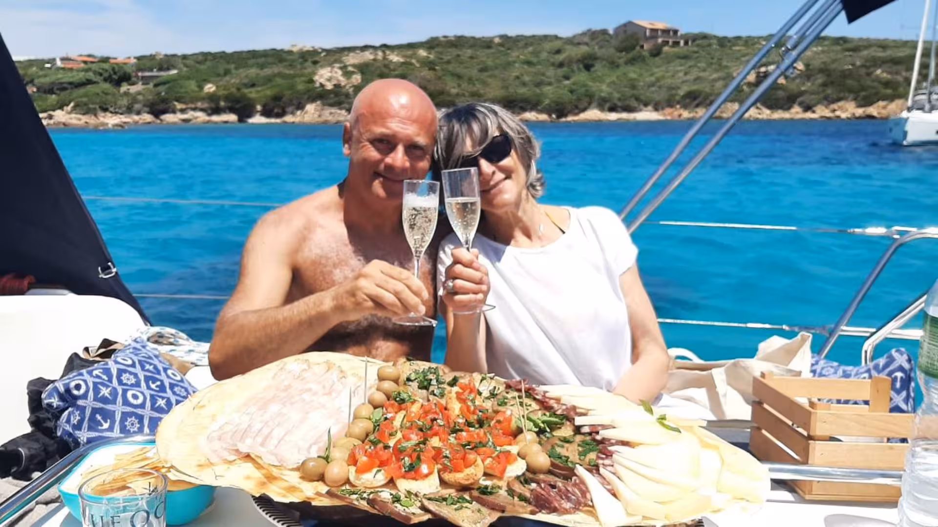 Couple toasting with champagne on a sailboat in La Maddalena Archipelago, featuring a delightful onboard lunch spread.