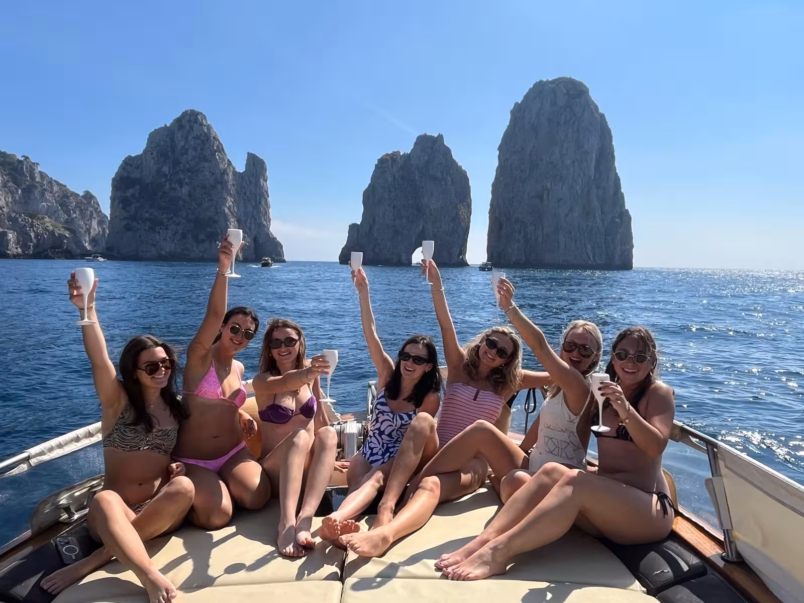 Smiling friends celebrating with drinks on a Capri boat tour, stunning Faraglioni rocks in the background.