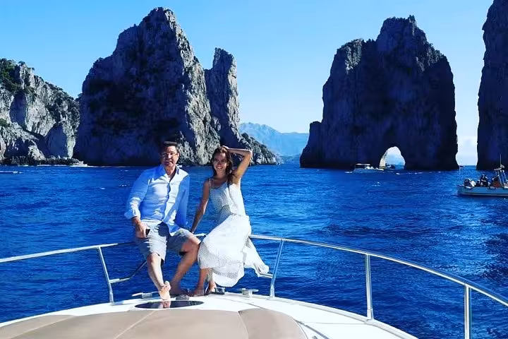 Couple on a Capri excursion boat posing with breathtaking Faraglioni sea stacks under a clear blue sky.