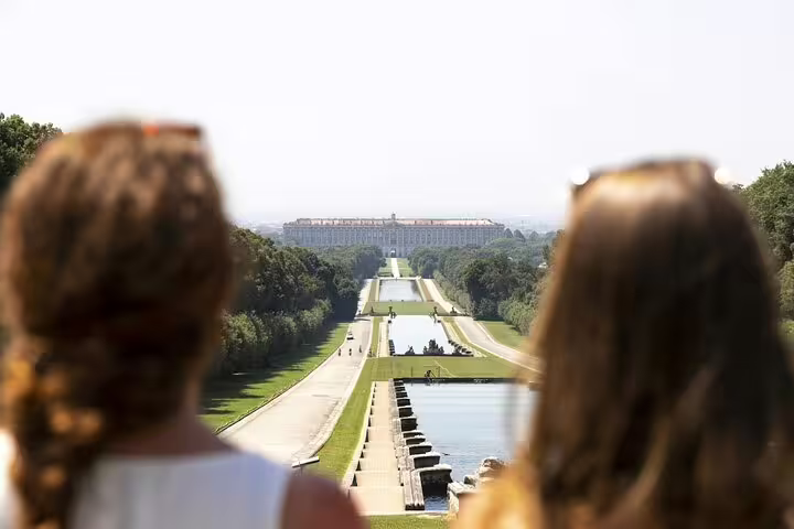 Two visitors admire the long fountains and tree-lined avenue leading to the Royal Palace of Caserta on a guided tour