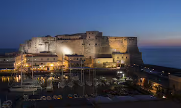 Castello Aragonese di Ischia illuminato al tramonto con porto turistico e barche a vela per la Festa di Sant’Anna in barca