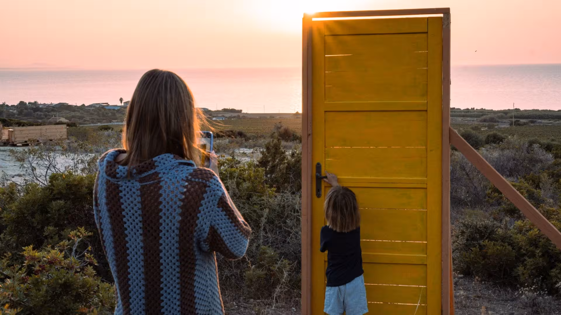 Woman and child near yellow door at sunset overlooking scenic Castelsardo landscape and ocean horizon.