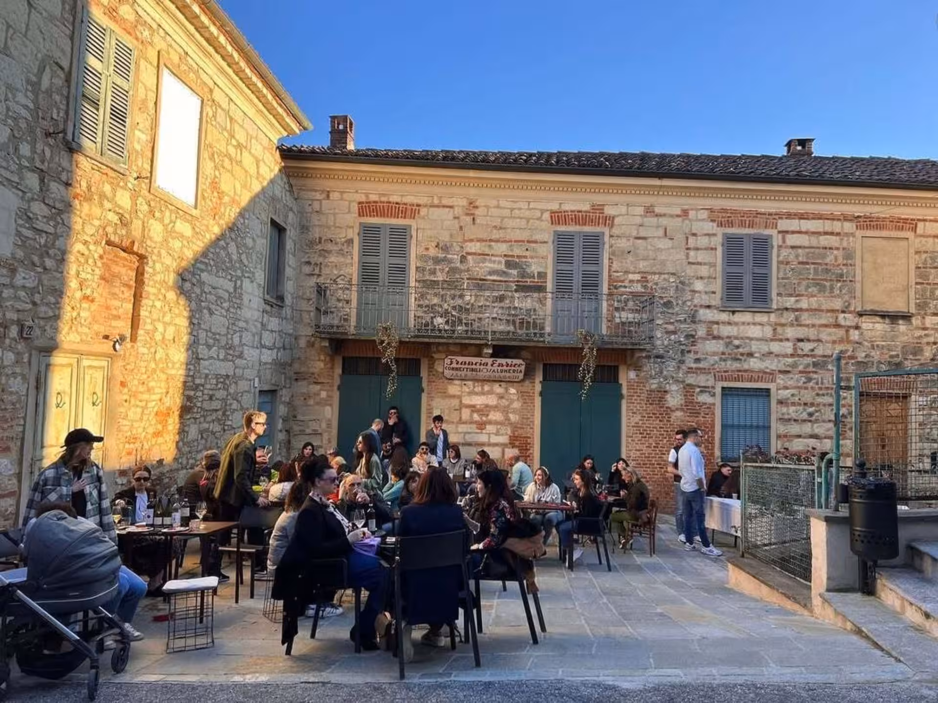 Visitors enjoying food and wine tasting in a sunlit rustic courtyard near Asti, creating a lively atmosphere.