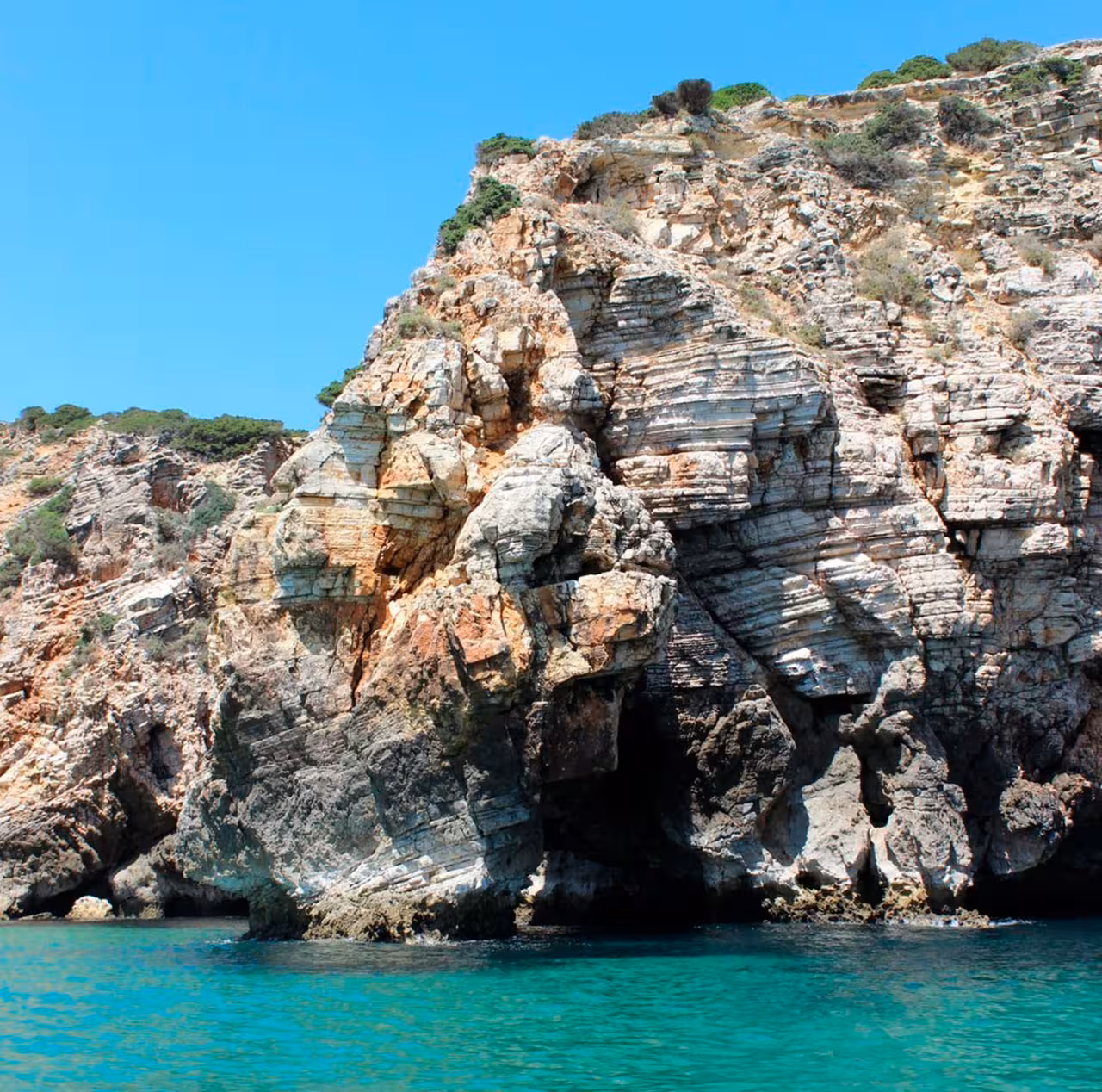 Majestic coastal cliff with rugged rock formations and crystal-clear turquoise water under a bright blue sky.