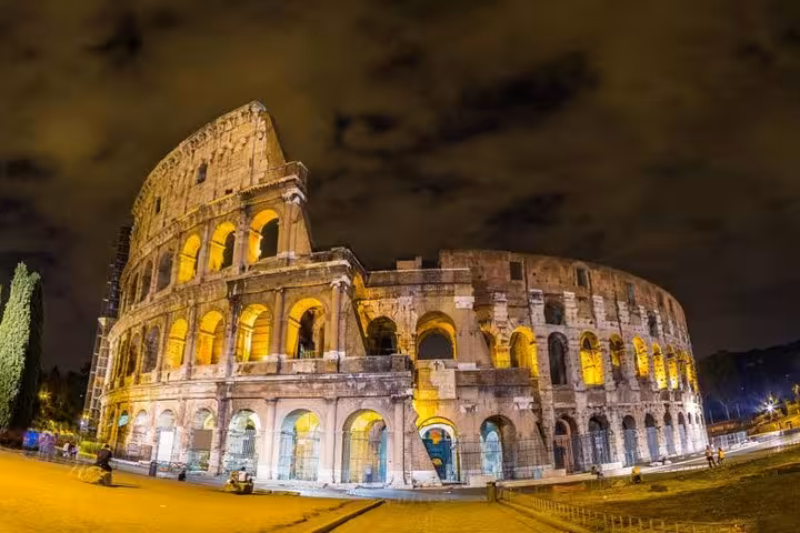 Illuminated Colosseum at night showcasing its grandeur against a dramatic sky, part of a guided night tour in Rome.