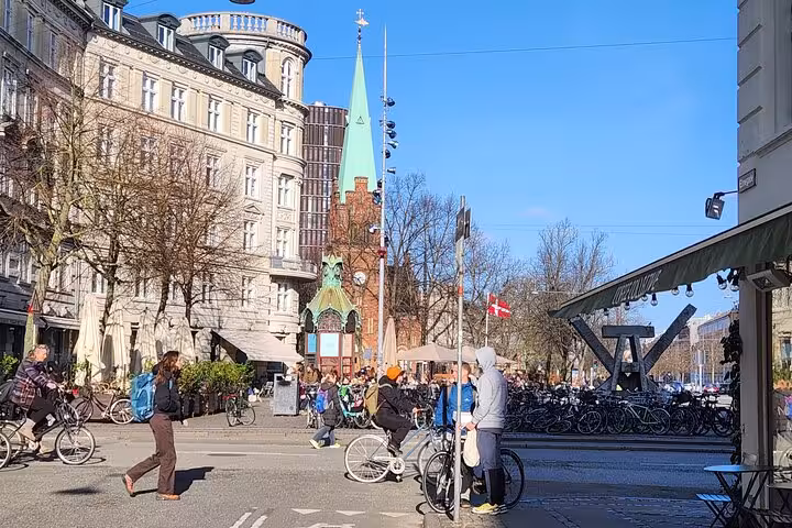 Cyclists and pedestrians enjoy a sunny day in central Copenhagen near historic architecture and Danish flags.