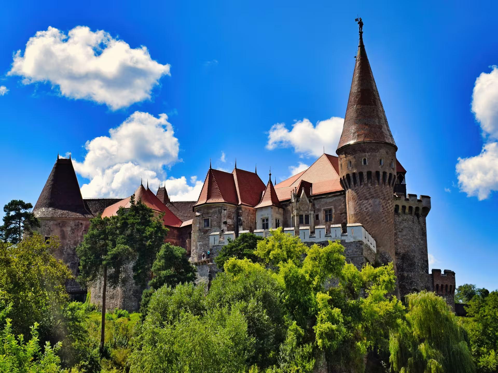 Stunning panoramic view of Corvin Castle in Transylvania surrounded by lush green trees under a bright blue sky
