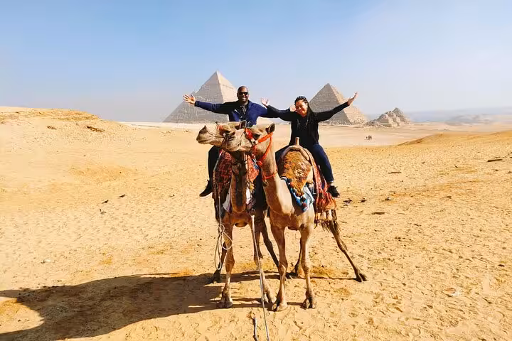 Couple posing on camels with the Giza Pyramids behind, private Giza tour with camel ride and ATV quad bike option