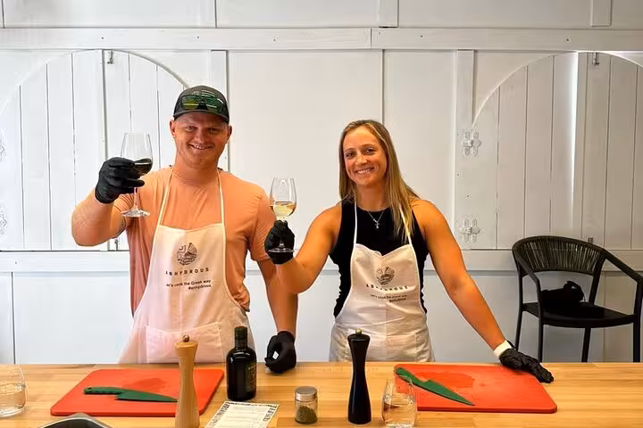 Couple toasting Santorini wine during honeymoon cooking class with aprons, prep station and local ingredients