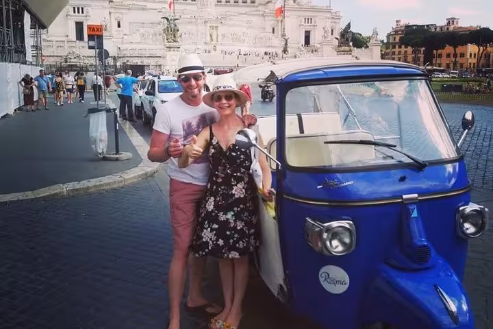 Couple enjoying a Valentine buggy tour in Rome, posing with a blue tuk-tuk near the historic Altare della Patria.
