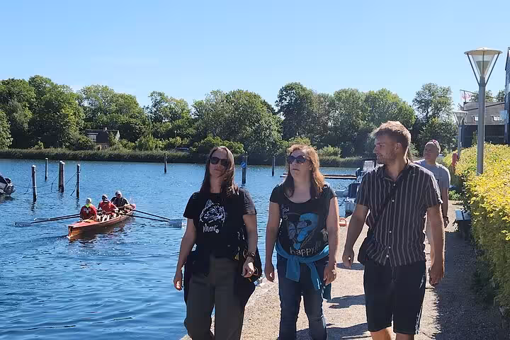 Group enjoying a leisurely walk by a scenic river with rowers, perfect for cultural and outdoor experiences.