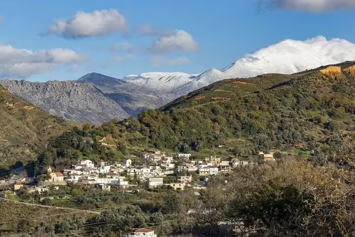 Scenic view of a picturesque village nestled in lush green hills with snow-capped mountains, ideal for a Heraklion wine tour.
