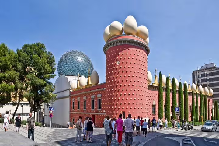 Visitors explore the iconic red facade of the Dalí Theatre-Museum adorned with large egg sculptures under a clear sky.