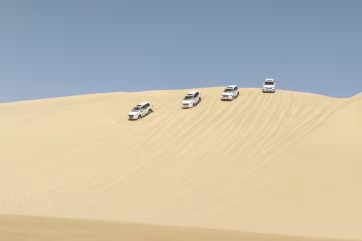 Four SUVs driving over a large sandy dune under a clear sky, showcasing an adventurous desert safari experience.