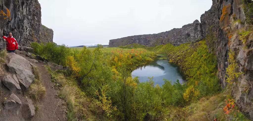 Hiker in red jacket overlooking a lush canyon with a small blue pond in Iceland