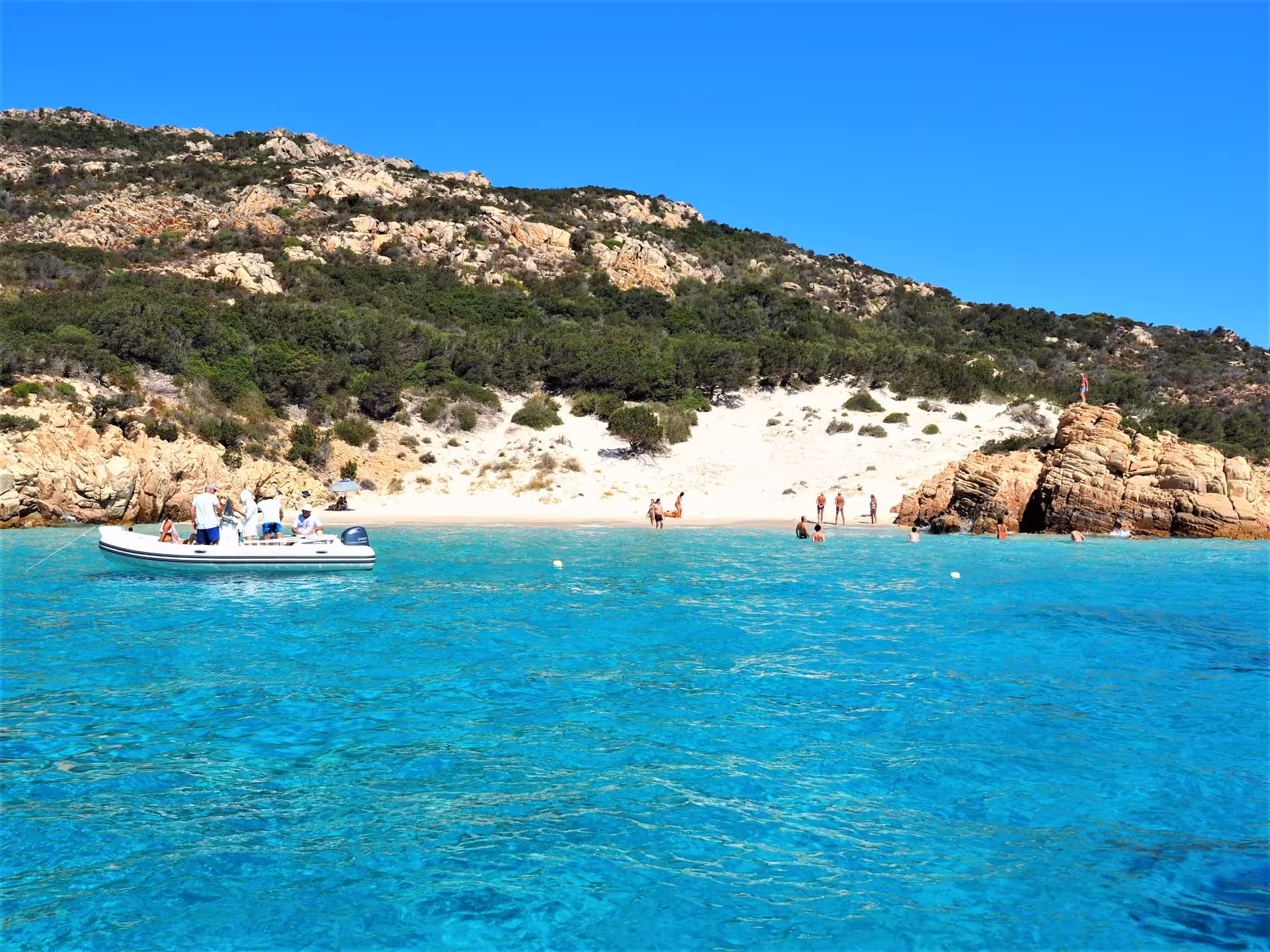 A dinghy anchored near the sandy shores of La Maddalena, with visitors enjoying the sun and stunning coastal views.
