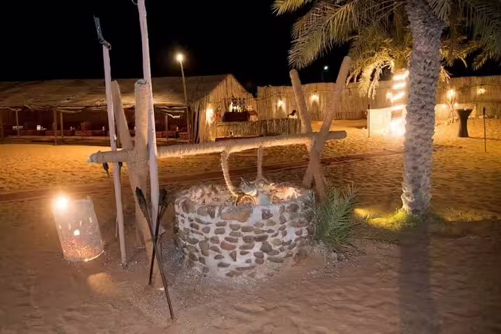 Traditional Bedouin desert camp at night in Dubai with fire pit, palm trees and seating for BBQ dinner safari guests