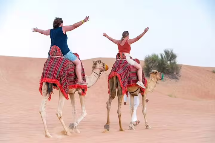 Two happy travelers riding camels across soft Dubai desert dunes, arms raised, on an adventurous evening desert safari tour
