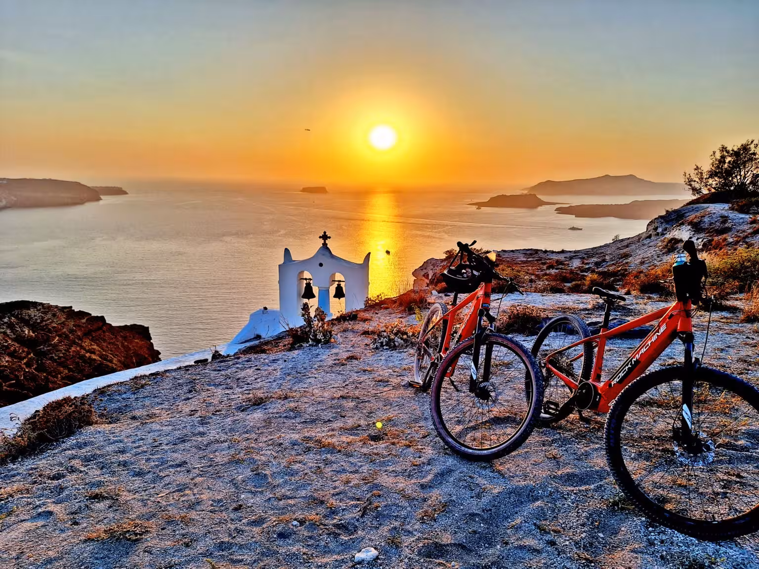 E-bike Santorini sunset tour with red electric bikes overlooking caldera and white chapel bell tower