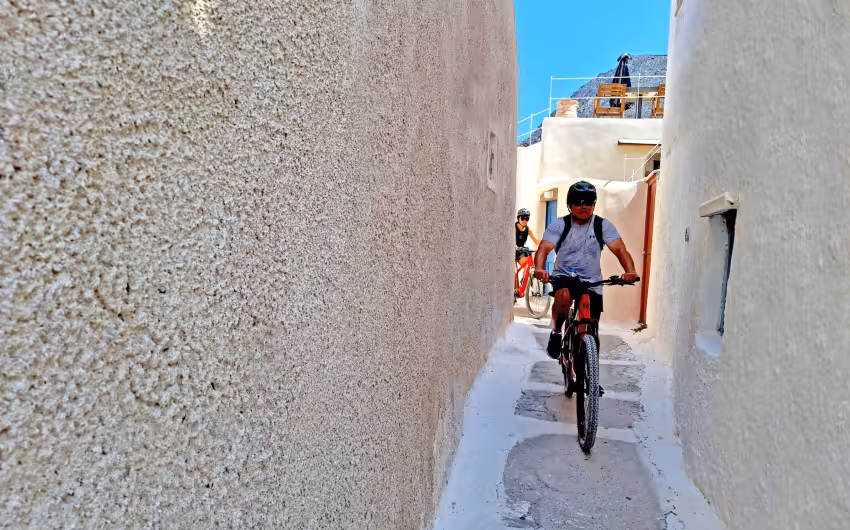 Riders on e-bikes cruise a narrow whitewashed alley in Santorini on a guided morning or sunset tour
