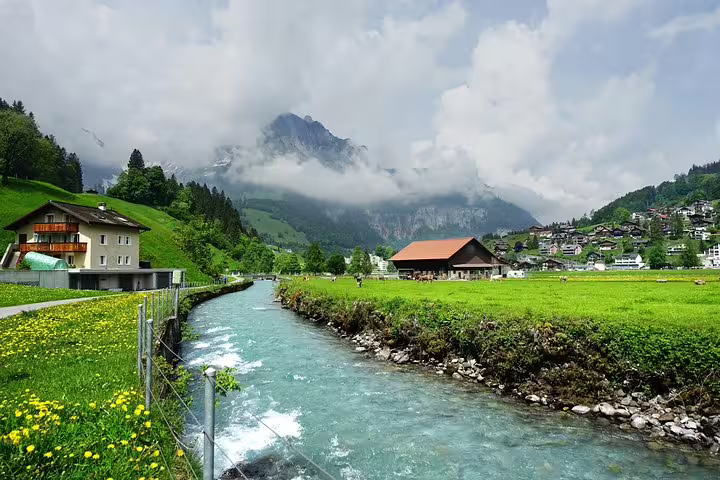 Scenic view of Engelberg with lush green fields, a flowing river, and charming houses against a backdrop of misty mountains.