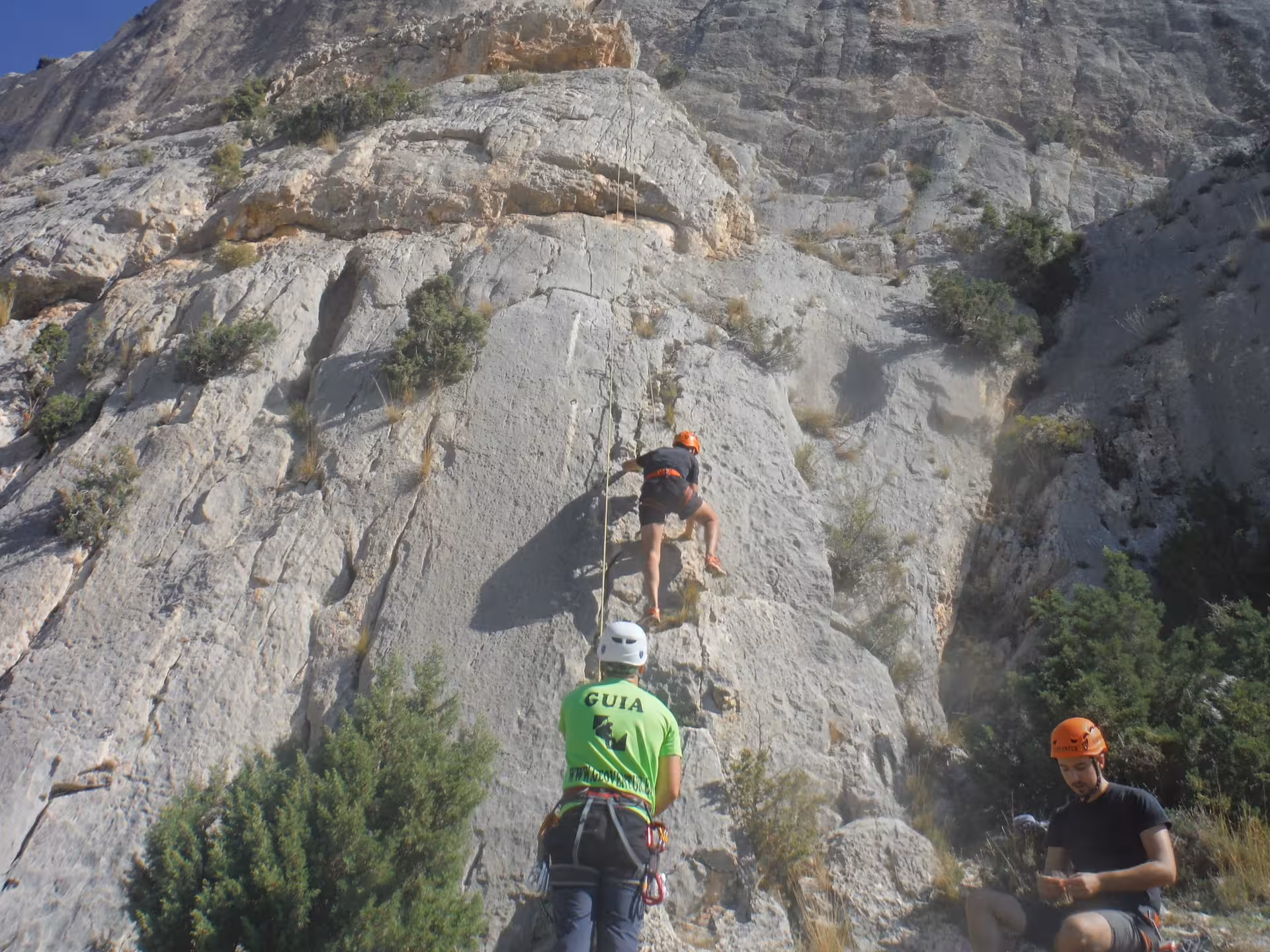 Guía de escalada asegura a principiante en vía de roca con cuerda y casco en curso de iniciación al aire libre