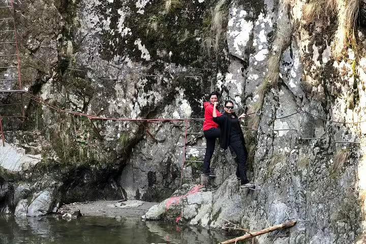 Two hikers posing on rocky cliffside trail with metal handrails along a river gorge