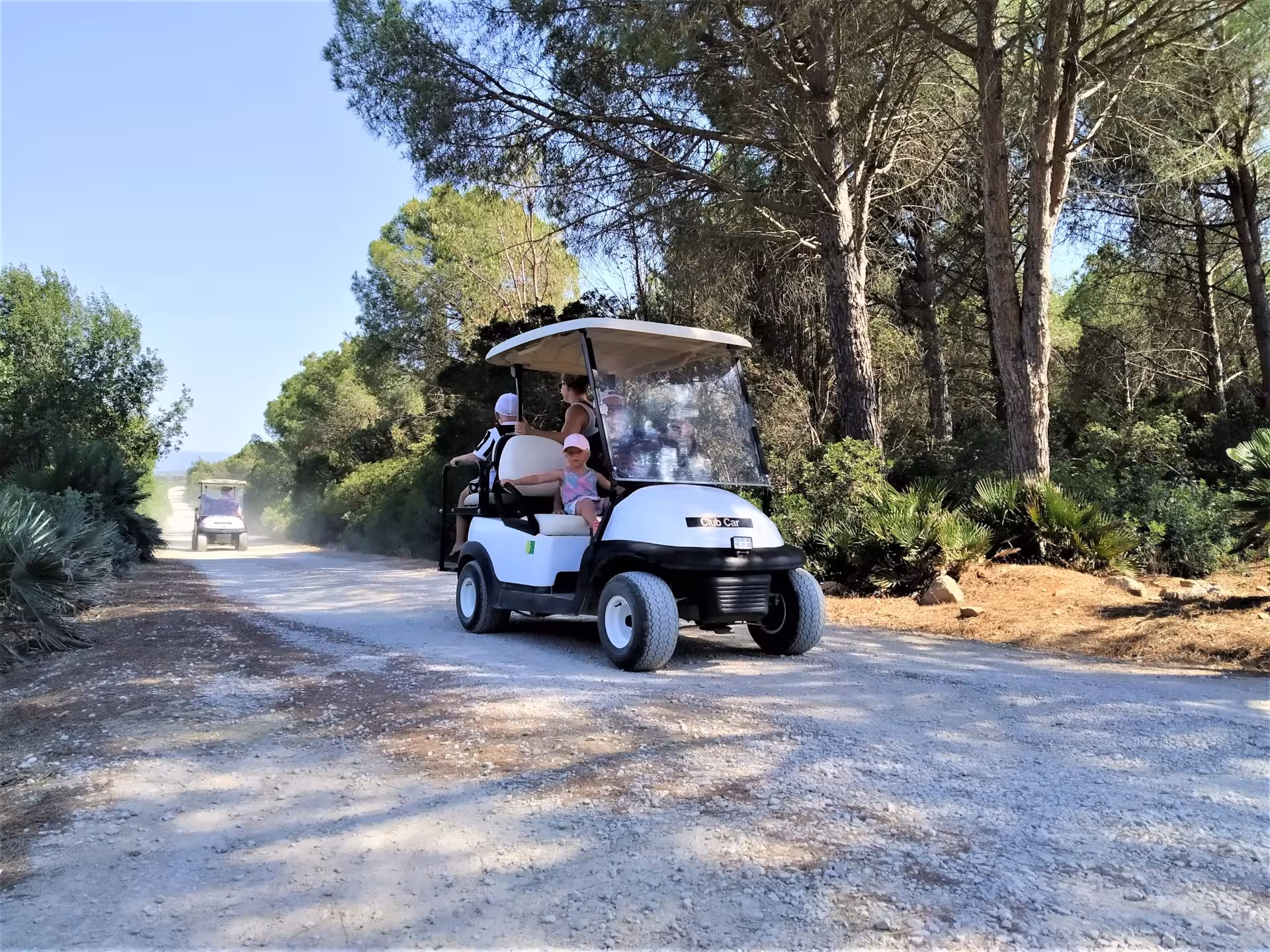 Family enjoying a golf cart ride on a scenic dirt path in Porto Conte Park, Alghero, surrounded by lush greenery.