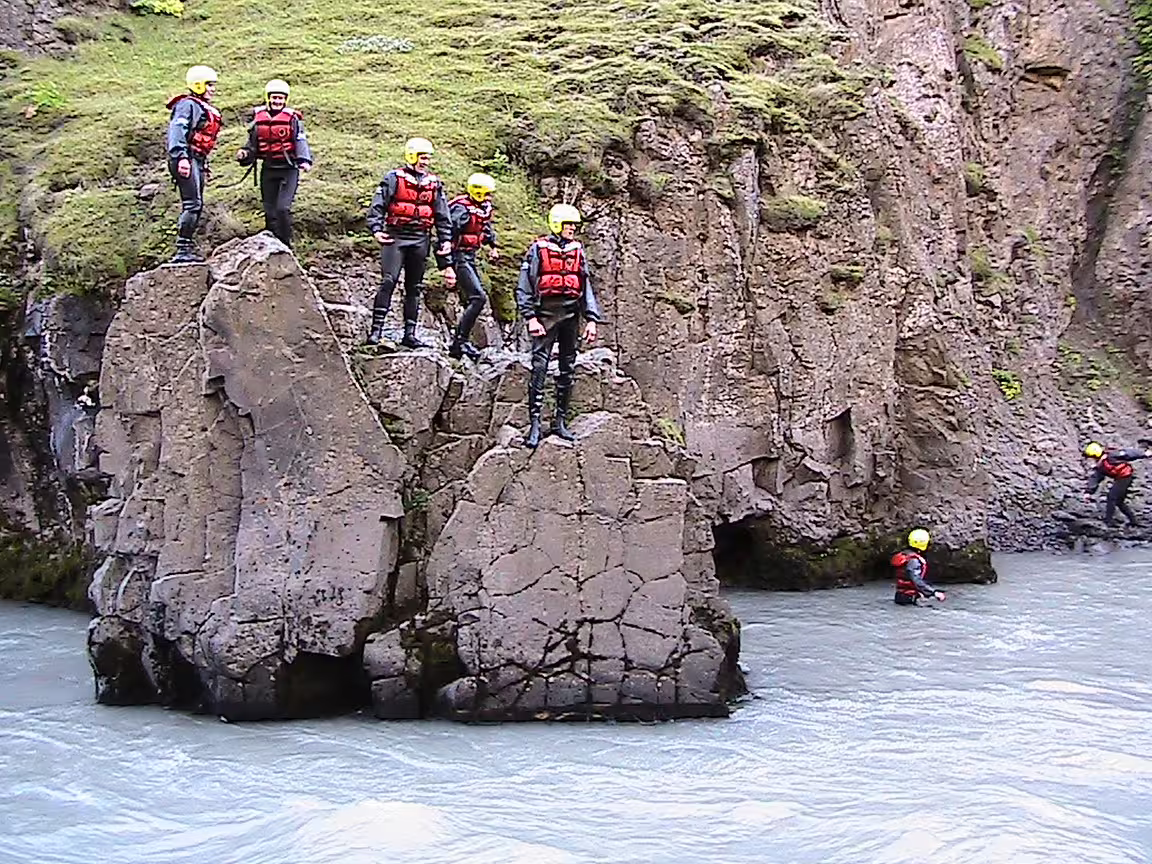 Family river rafting adventure with group preparing to launch into rapids, wearing safety gear, surrounded by scenic cliffs.