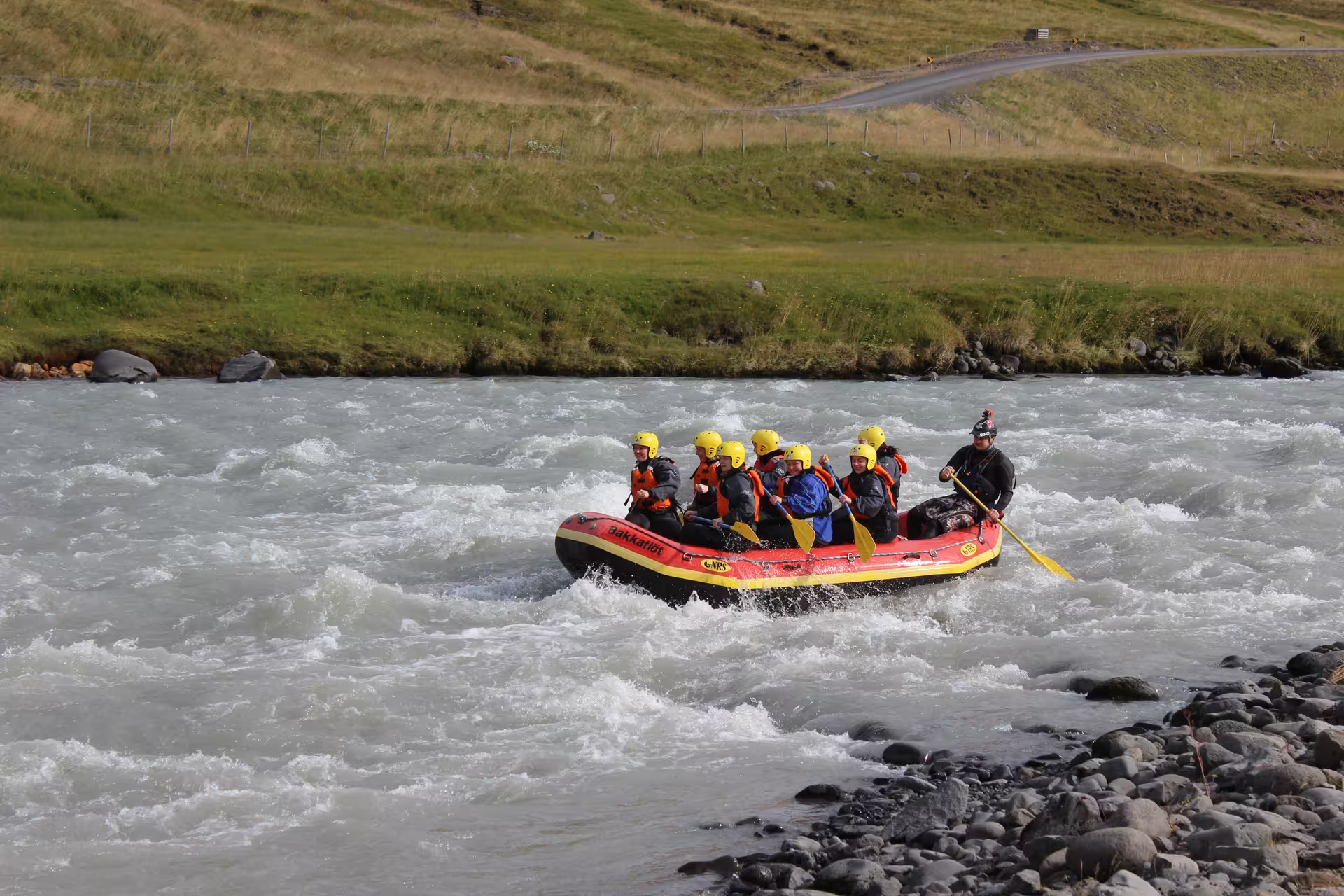 Families enjoy thrilling river rafting adventure on a scenic, rocky river with lush green hills in the background.