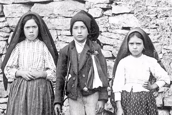Three children in traditional attire stand against a stone wall, representing the historical and cultural heritage of Fatima.