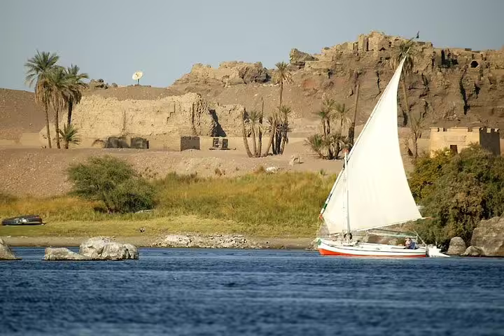 Traditional felucca boat sailing the Nile in Luxor past palm trees and ancient ruins on the West Bank