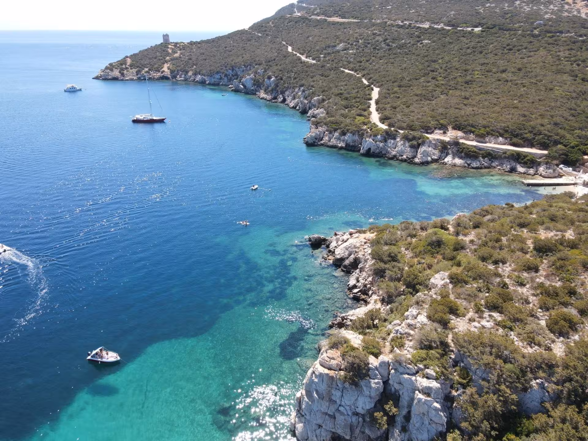 Aerial view of boats sailing in the crystal-clear waters of the Gulf of Alghero, surrounded by lush coastal cliffs.