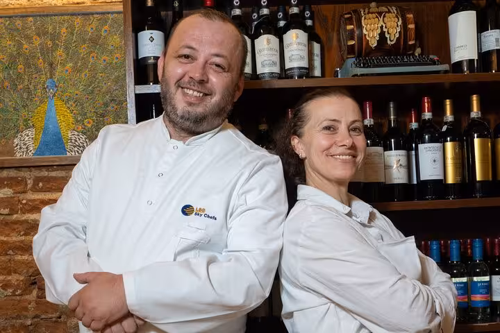 Smiling chefs in Florence posing in front of a wine display, ready for a pasta and tiramisu cooking class.