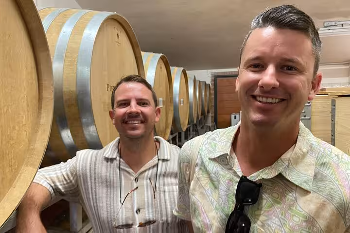 Smiling friends on a wine tasting tour stand beside large wooden barrels in a Tuscan winery.