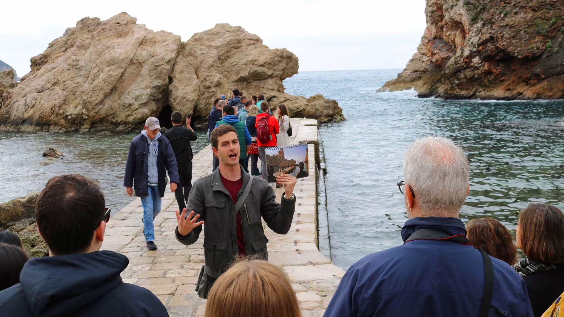 Guide leads Game of Thrones and Dubrovnik history group tour on seaside pier by rocky cliffs and Adriatic