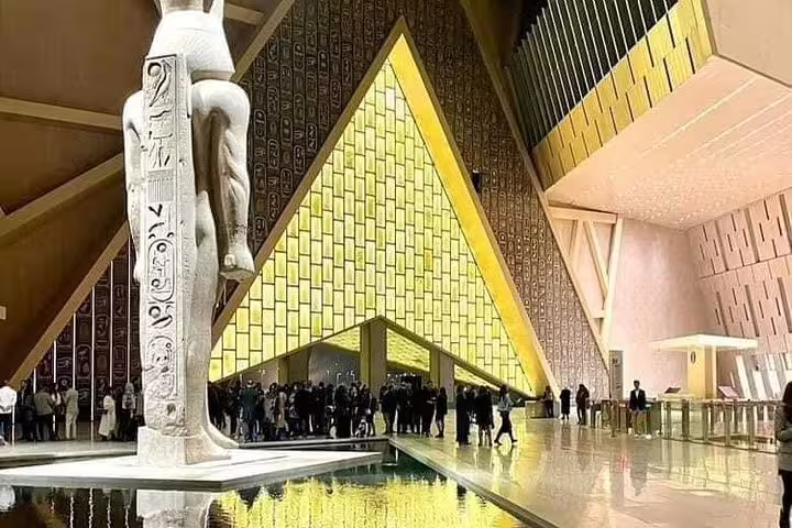 Visitors gather at Grand Egyptian Museum GEM atrium by colossal statue and illuminated pyramid wall on 3-hour tour