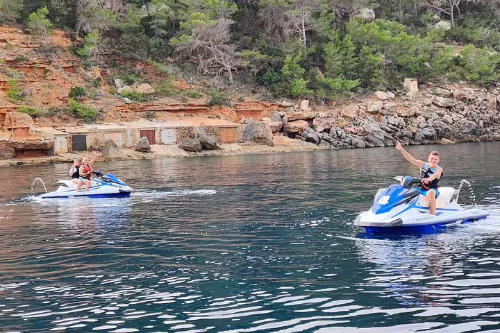 Friends on jet skis during guided Cala Salada Ibiza excursion, waving on calm water by rocky shoreline