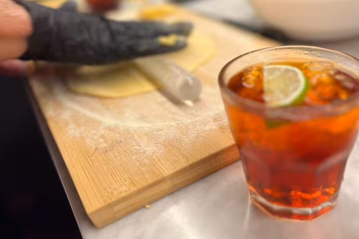 Close-up of a hand rolling pasta dough on a wooden board beside a refreshing spritz cocktail with lime.