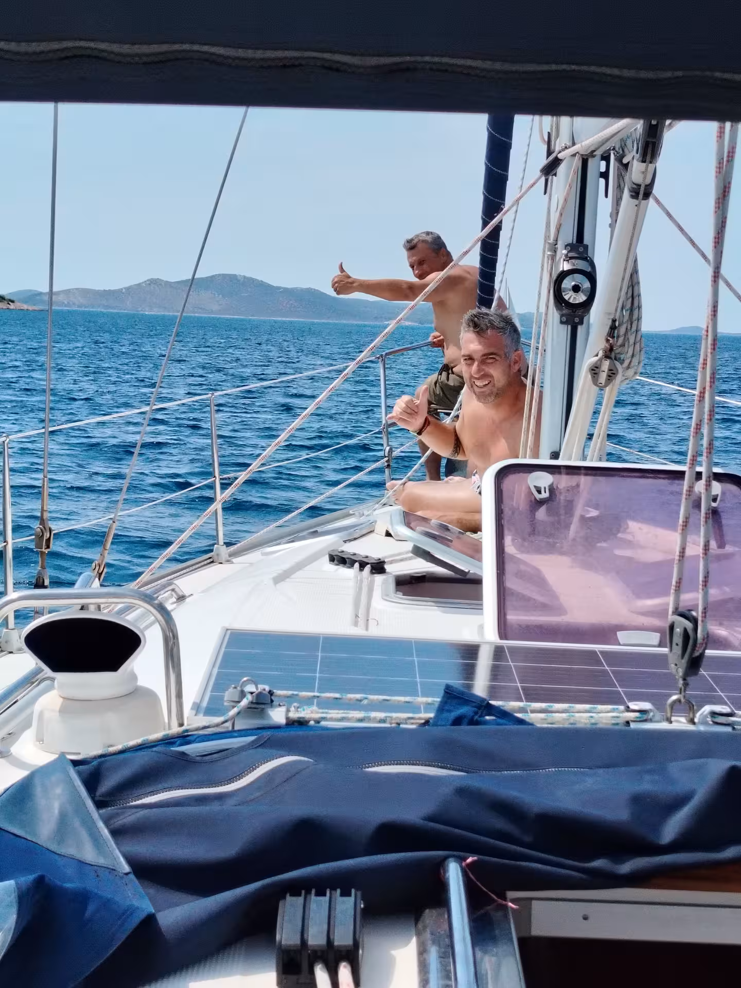 Happy guests on sailboat deck giving thumbs up during coastal sailing adventure, crystal-clear sea and islands