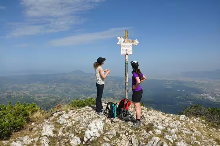 Two hikers at a trail signpost on a rocky mountain summit with panoramic valley views