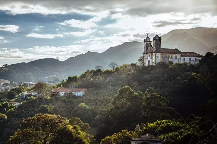 Scenic view of a historic church perched on a lush hillside in Minas Gerais, Brazil, under a dramatic sky.