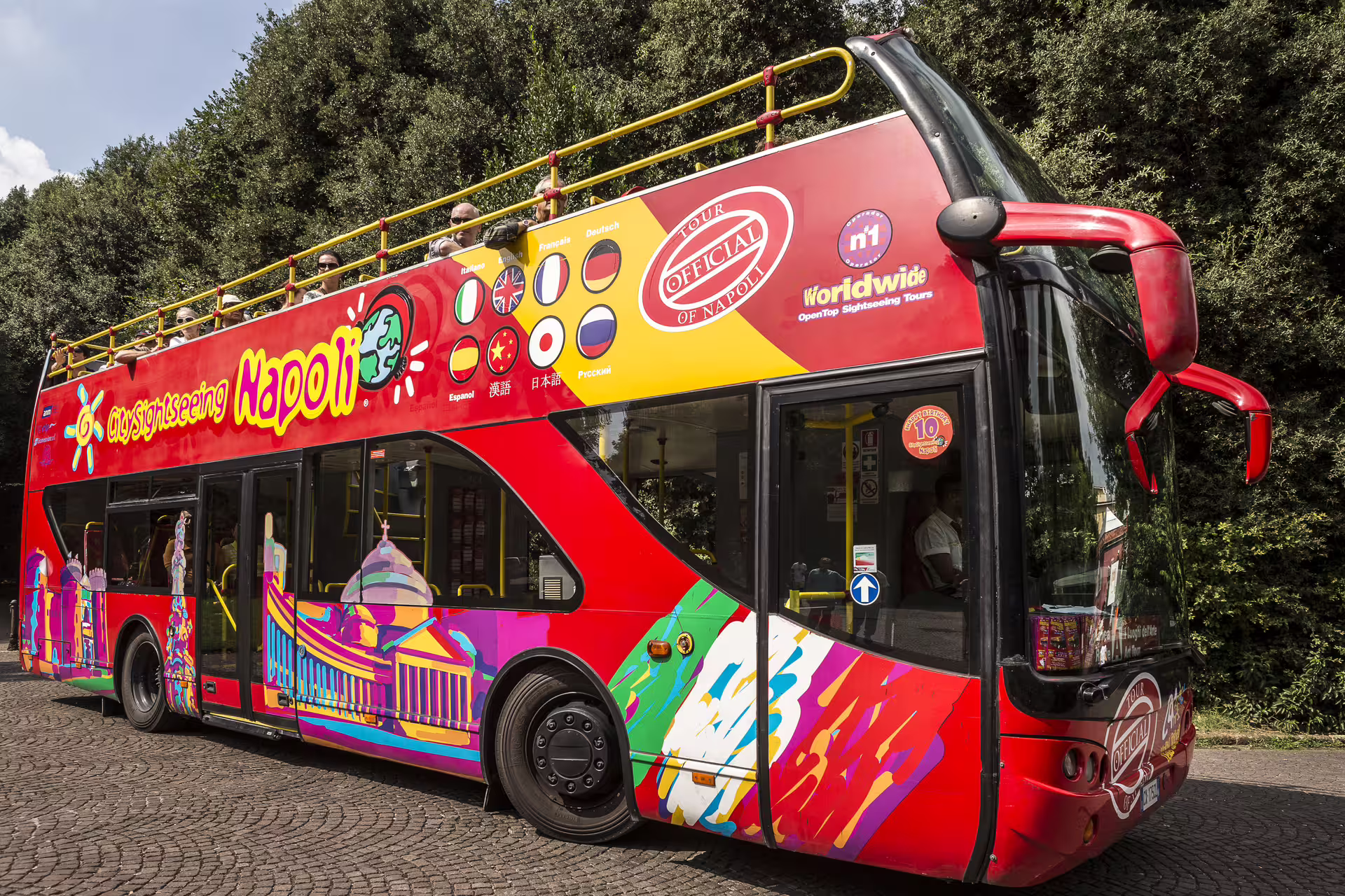Colorful City Sightseeing Napoli hop on hop off bus waiting under trees, ready for panoramic tours of Naples attractions