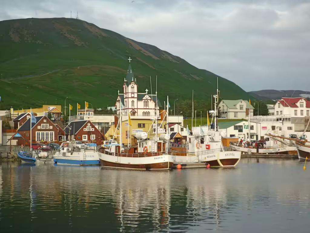 Husavik harbor with colorful boats, historic church, and green mountain backdrop on Diamond Circle tour