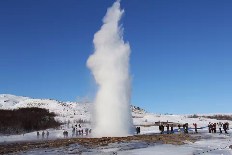Tourists admire a powerful geyser eruption in the snowy landscape of Iceland's Golden Circle under a clear blue sky.