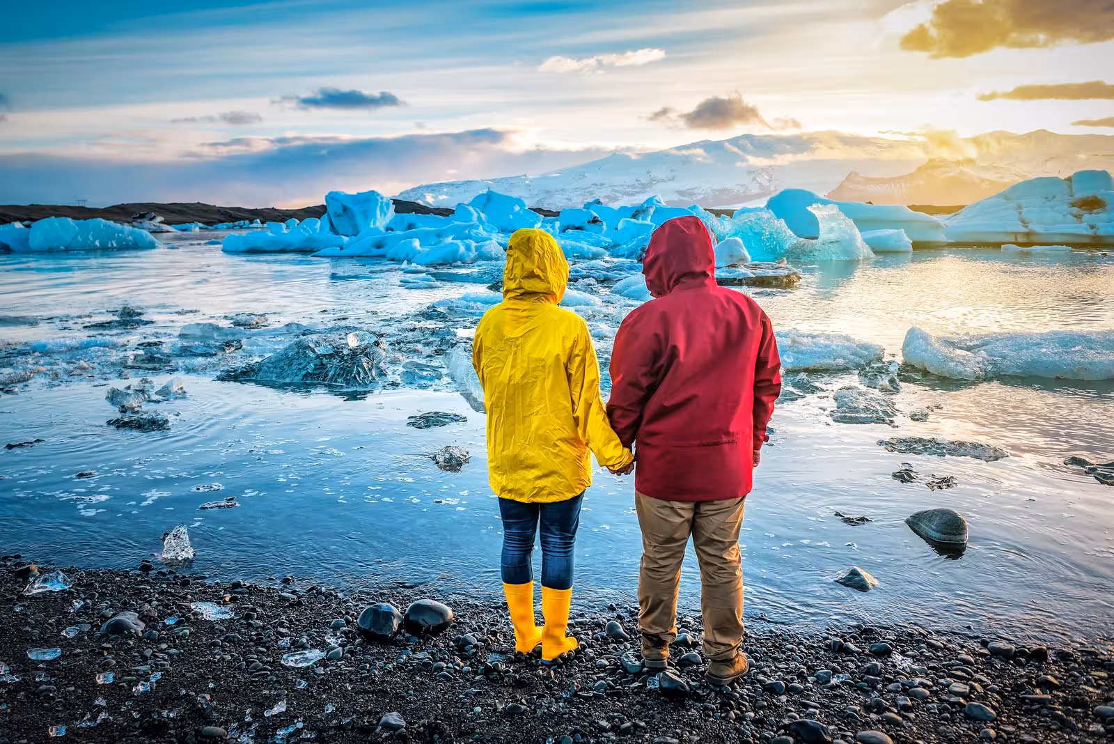 Couple in vibrant jackets holding hands at Iceland's Jokulsarlon Glacier Lagoon during sunset on the Grand Tour.